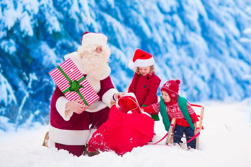 Santa Claus and children opening presents in snowy forest. Kids and father in Santa costume and beard open Christmas gifts. Little girl helping with present sack. Xmas, snow and winter fun for family.