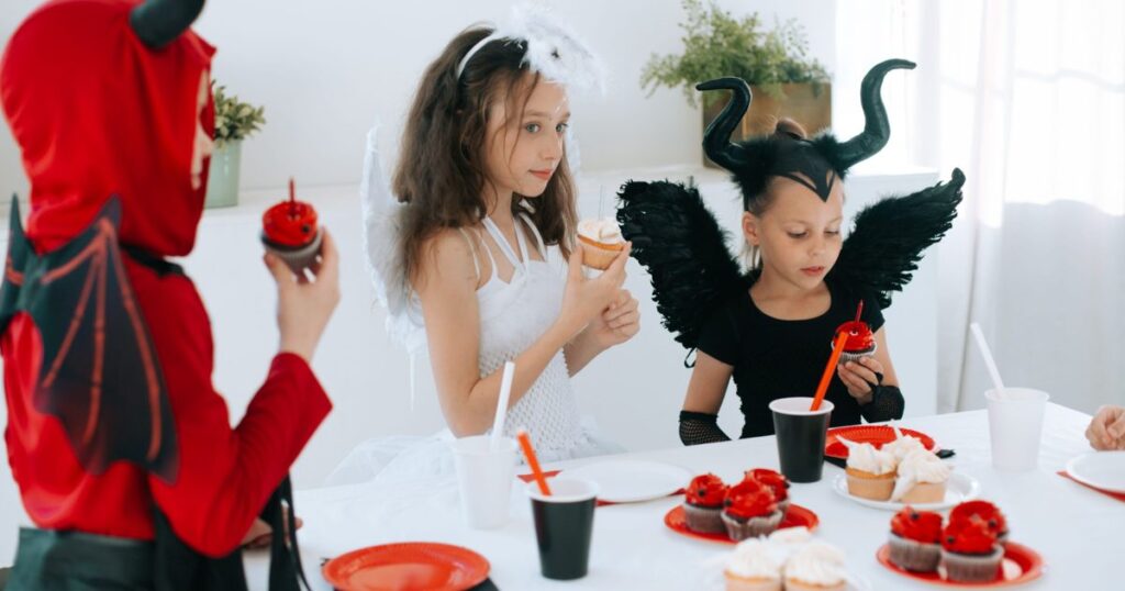 Tres niños disfrazados de diablo, ángel y criatura con cuernos merendando cupcakes en una fiesta de Halloween