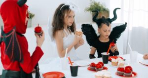 Tres niños disfrazados de diablo, ángel y criatura con cuernos merendando cupcakes en una fiesta de Halloween