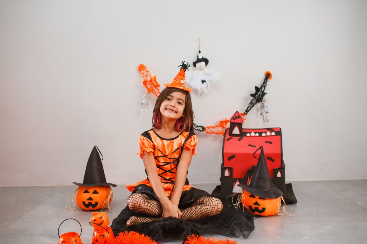 Niña con vestido de bruja naranja y negro y sombrero pequeño con araña, sentada entre calabazas