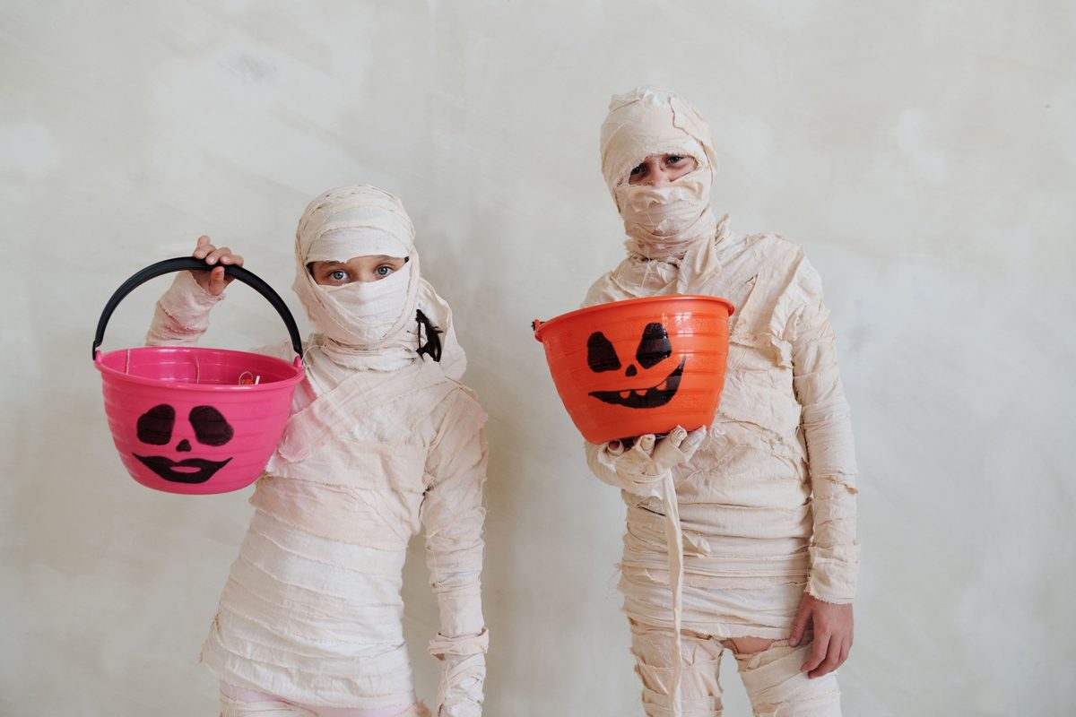 Dos niños disfrazados de momia con tiras de tela beis y cubos de calabaza para los caramelos