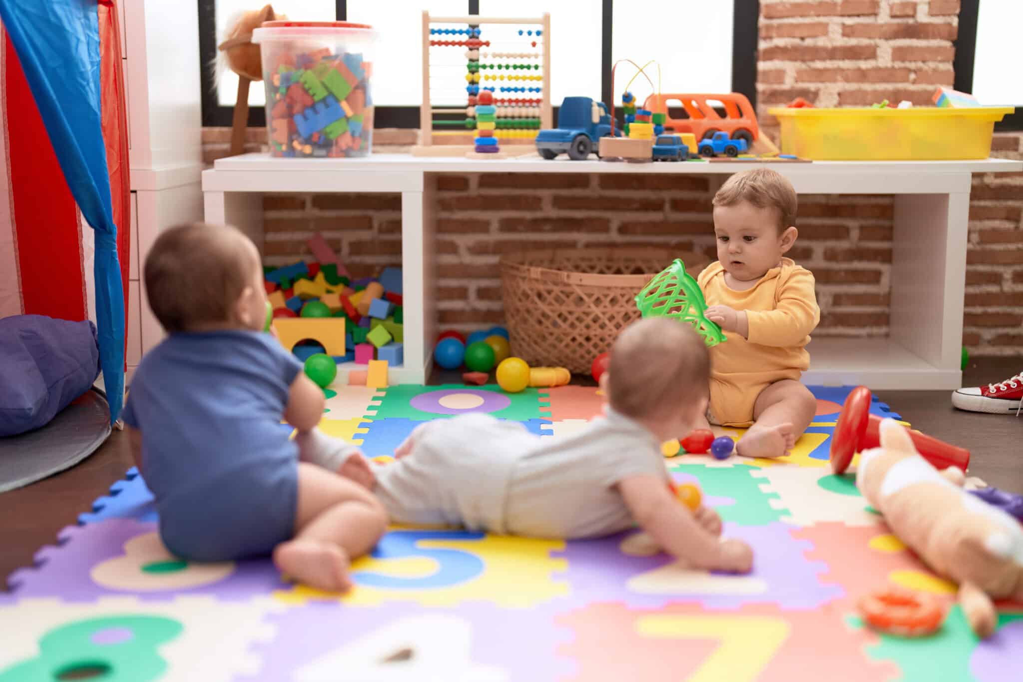 Tres bebés jugando en una alfombra de puzle con juguetes y bloques de colores en una escuela infantil