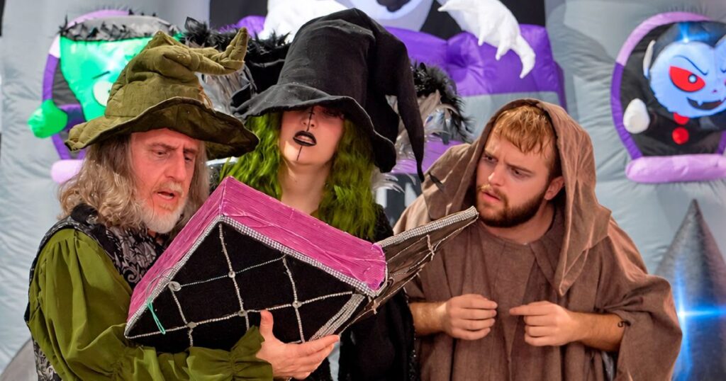 Actores disfrazados de mago, bruja y monje leen un libro gigante en el Halloween del Centro Comercial Arena